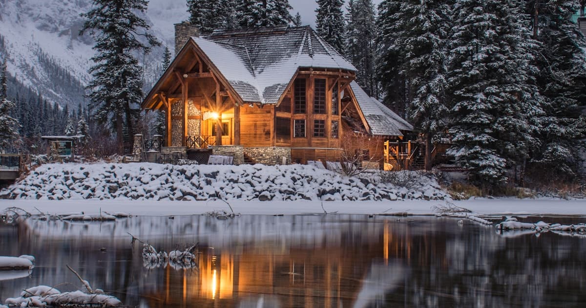 Chalet d'hiver au bord d'un lac avec une lumière chaleureuse aux fenêtres, entouré d'arbres enneigés